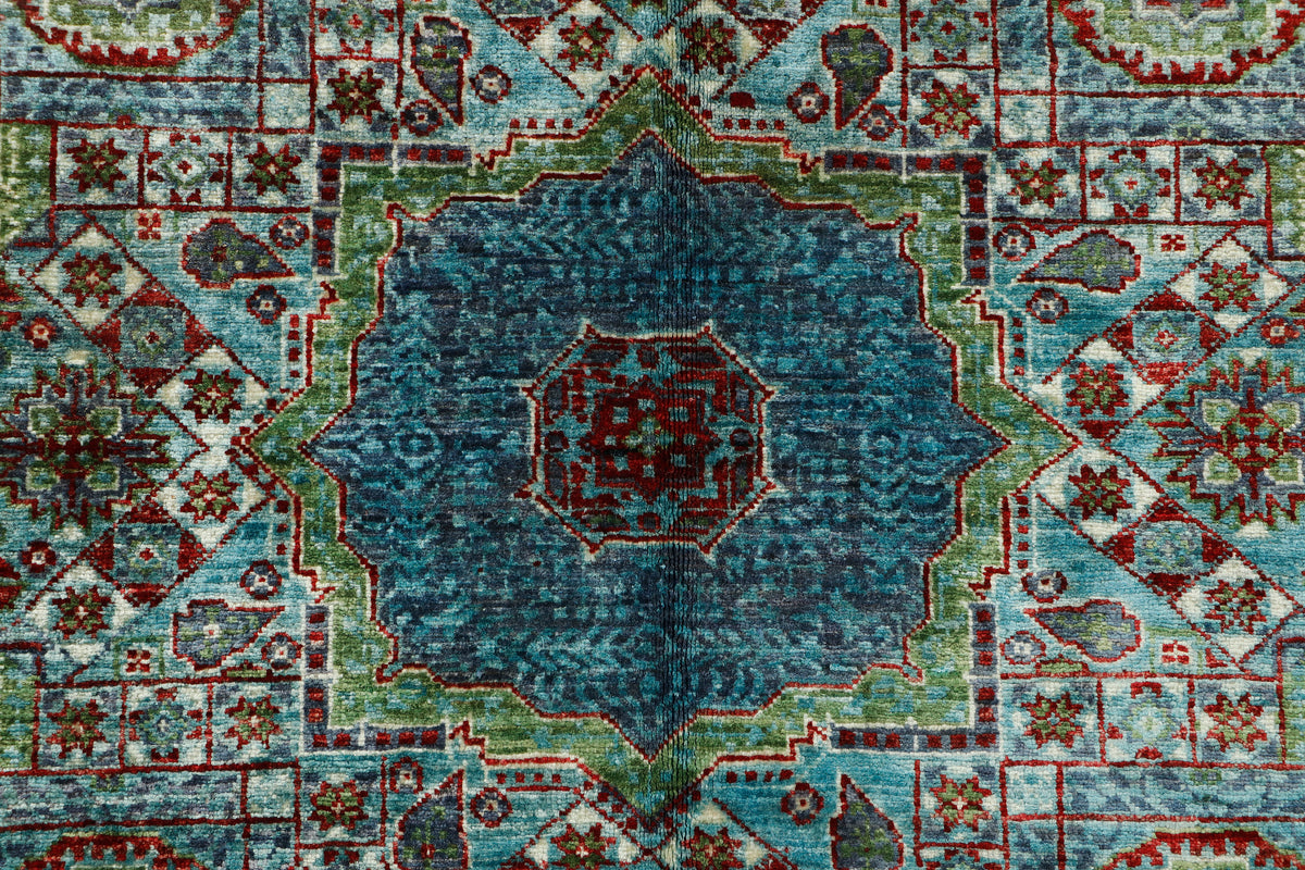 A close-up of a traditional patterned rug featuring a central medallion in shades of blue and teal, surrounded by green and red geometric motifs on a light blue field.