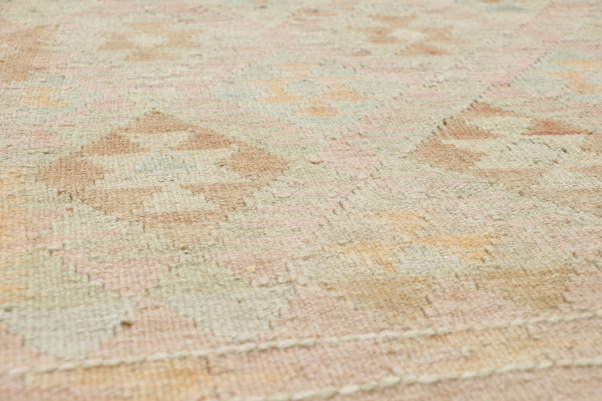 A close-up of a woven rug with a soft, textured surface featuring a geometric diamond pattern in muted pastel shades of pink, beige, and light green.