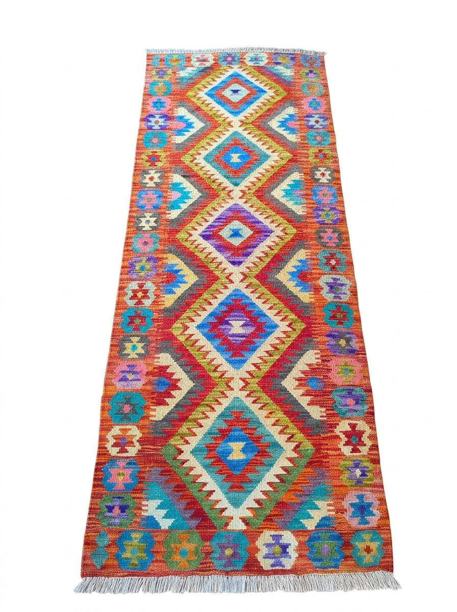 A long rectangular woven runner rug with a bold geometric diamond pattern in red, orange, blue, teal, cream, and purple, finished with white fringe on the ends.